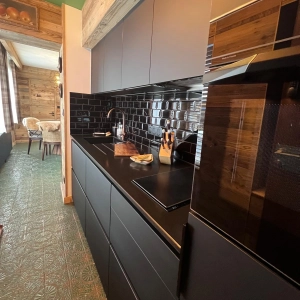 Cuisine de chalet de montagne rénovée, meubles noirs mats, plan de travail noir, crédence bordeaux avec joints pailletés, cuisine aménagée sur mesure